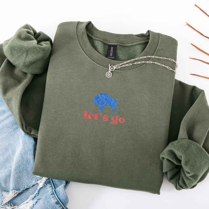 May include: An olive green sweatshirt with a blue bison graphic and the words "let's go" in red. A silver necklace with a circular pendant is on the sweatshirt. The sweatshirt is folded with a pair of blue jeans.