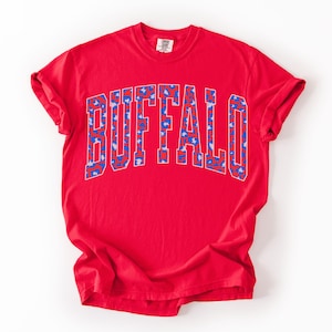 May include: A red t-shirt with the word "BUFFALO" printed across the chest in a blue and white leopard print design. The shirt has short sleeves and a crew neck.
