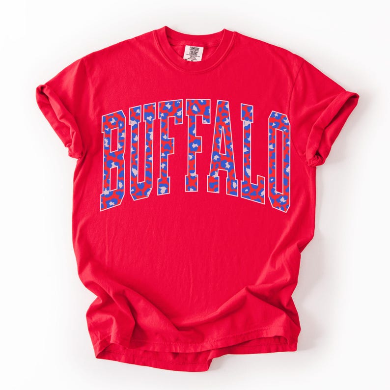 May include: A red t-shirt with the word "BUFFALO" printed across the chest in a blue and white leopard print design. The shirt has short sleeves and a crew neck.