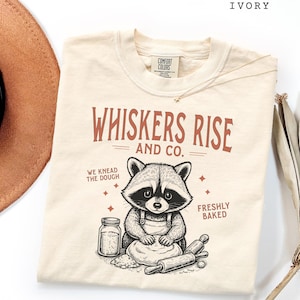 May include: Ivory t-shirt featuring the text "WHISKERS RISE AND CO." and a raccoon graphic. The raccoon is wearing an apron and kneading dough. The shirt also features the phrases "WE KNEAD THE DOUGH" and "FRESHLY BAKED".