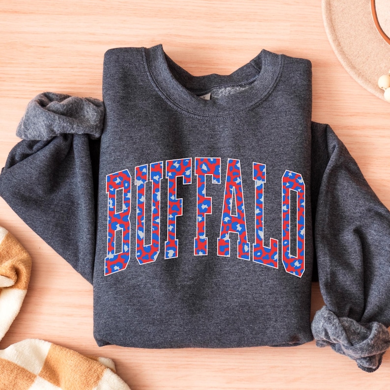 May include: A dark gray sweatshirt with the word "BUFFALO" in large, block letters across the chest. The letters are filled with a red, white, and blue pattern. The sweatshirt is folded on a light wooden surface.