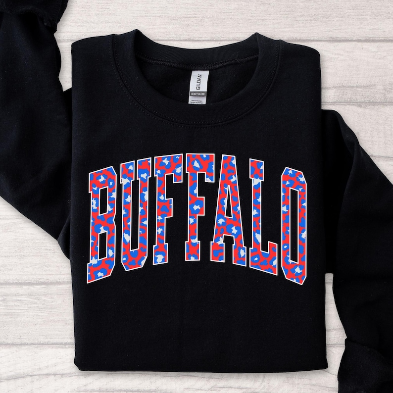 A black sweatshirt featuring bold arched “BUFFALO” varsity lettering filled with a bright red-and-blue patterned print and outlined in white. The vibrant design stands out sharply against the dark fabric, giving it a trendy, sporty Buffalo-pride look.