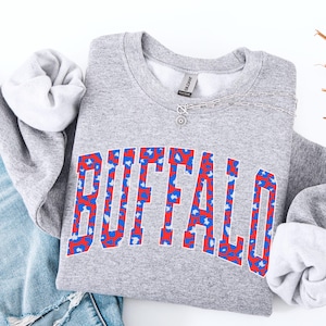 May include: A heather gray sweatshirt with the word "BUFFALO" in large, bold letters across the front. The letters are outlined in red and filled with a blue and red leopard print pattern. A silver necklace with a small pendant is draped across the sweatshirt.