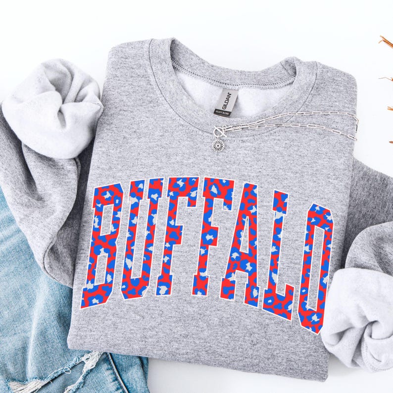 May include: A heather gray sweatshirt with the word "BUFFALO" in large, bold letters across the front. The letters are outlined in red and filled with a blue and red leopard print pattern. A silver necklace with a small pendant is draped across the sweatshirt.
