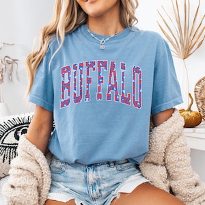 May include: A light blue t-shirt with the word "BUFFALO" in a red, white, and blue leopard print design. The shirt has a crew neck and short sleeves. The shirt is made of a soft material.