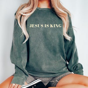 May include: A dark green sweatshirt with the text "JESUS IS KING" in a light beige color. The long-sleeved top has a slightly faded appearance. The sweatshirt is a casual style.