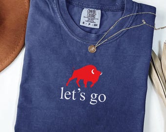 May include: A navy blue t-shirt featuring a red buffalo graphic and the words "let's go" in white. A gold necklace with a pendant rests on the shirt. The shirt is folded and has a Comfort Colors label.