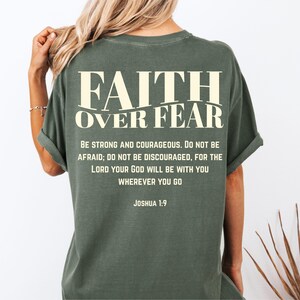 May include: Olive green t-shirt with the text "FAITH OVER FEAR" in a cream-colored font. Below the text is a quote from Joshua 1:9, encouraging strength and courage. The shirt has rolled-up sleeves.