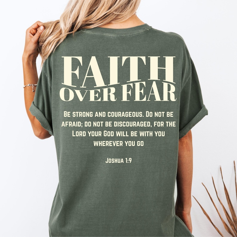 May include: Olive green t-shirt with the text "FAITH OVER FEAR" in a cream-colored font. Below the text is a quote from Joshua 1:9, encouraging strength and courage. The shirt has rolled-up sleeves.