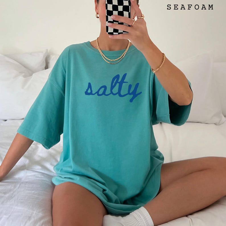以下が含まれることがあります： 前面に青い筆記体で「salty」と書かれた、ターコイズ色のTシャツ。 ゆったりとしたフィット感の半袖シャツです。 右上には白で「SEAFOAM」の文字がプリントされています。