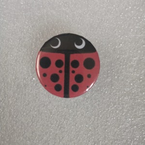 May include: A red ladybug pin with black spots and white eyes. The pin is round and has a shiny finish.