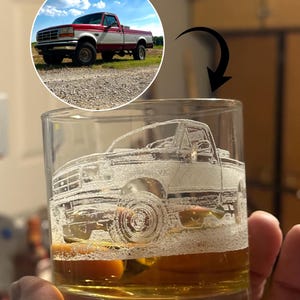 May include: A clear glass tumbler etched with a detailed image of a red and white pickup truck. The glass is partially filled with a golden-colored liquid. A circular image of the truck is above the glass, showing the vehicle on a gravel road under a blue sky.