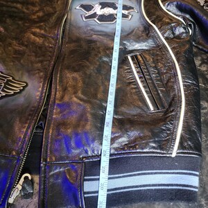 May include: A black leather jacket with white piping, a front zipper, and ribbed cuffs. A measuring tape is placed vertically along the front, showing a length of approximately 33 inches. The jacket features embroidered patches and a collar.