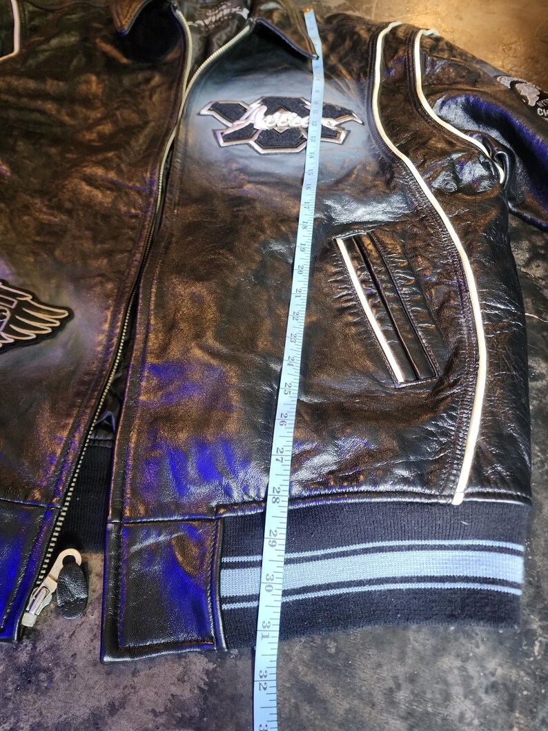 May include: A black leather jacket with white piping, a front zipper, and ribbed cuffs. A measuring tape is placed vertically along the front, showing a length of approximately 33 inches. The jacket features embroidered patches and a collar.