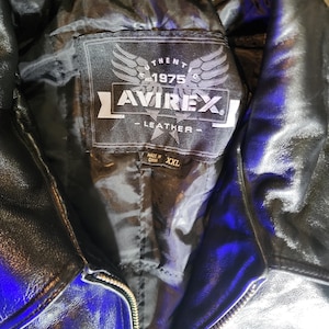 May include: A black leather jacket with a black interior lining. The inner label features the text AVIREX, 1975, and LEATHER with a wing graphic. A smaller tag below the main label indicates the size as XXL and states Made in China.