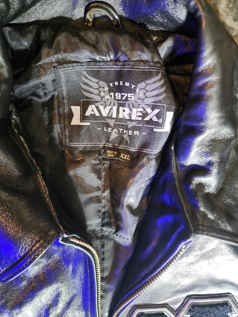 May include: A black leather jacket with a black interior lining. The inner label features the text AVIREX, 1975, and LEATHER with a wing graphic. A smaller tag below the main label indicates the size as XXL and states Made in China.
