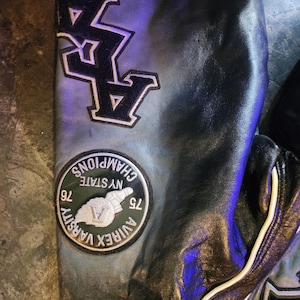 May include: A black leather jacket sleeve featuring a large black and white embroidered patch with the text AVIREX VARSITY CHAMPIONS NY STATE 76 75 and a large black letter A patch. The leather has a smooth texture with visible white piping.