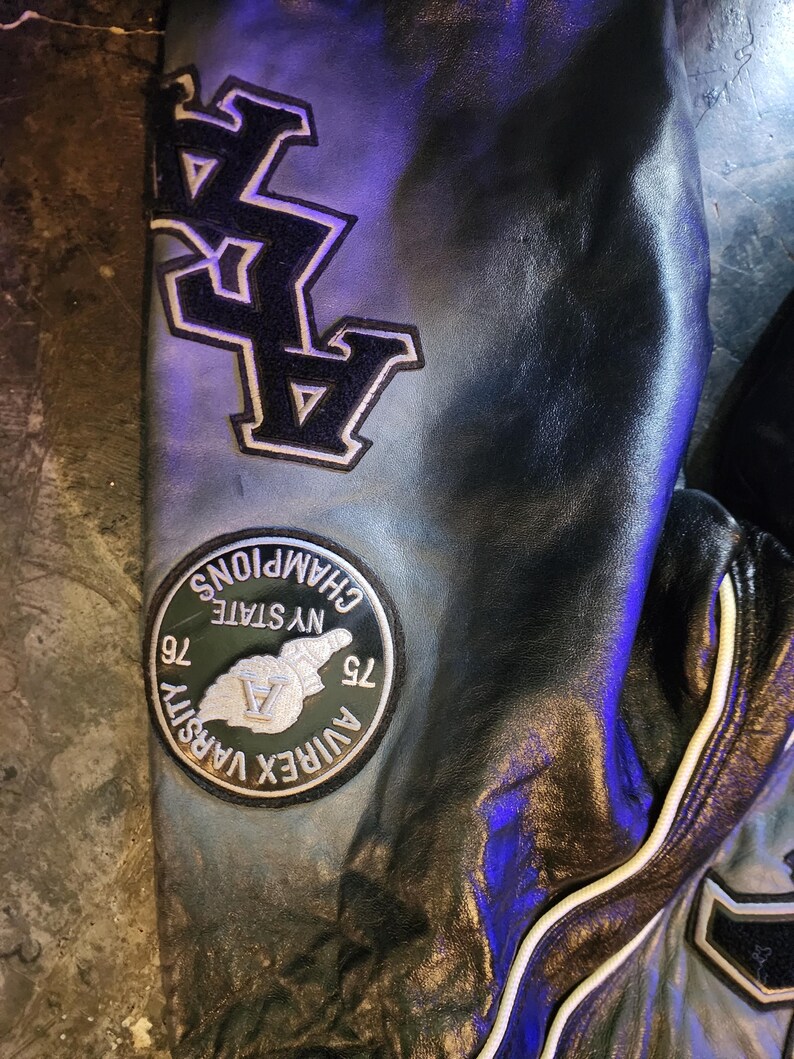 May include: A black leather jacket sleeve featuring a large black and white embroidered patch with the text AVIREX VARSITY CHAMPIONS NY STATE 76 75 and a large black letter A patch. The leather has a smooth texture with visible white piping.
