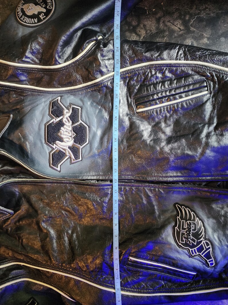 May include: A black leather jacket with white piping, featuring embroidered patches including the text 'Avirex' and '75'. A blue measuring tape is placed vertically over the center, showing a length of approximately 27 inches from the top to the hem.