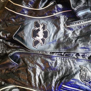 May include: A black leather jacket featuring white piping, a front zipper closure, and embroidered patches. The chest displays a large Avirex logo patch, while the lower section includes a winged emblem. The interior label reads Avirex 1875.