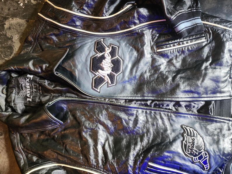 May include: A black leather jacket featuring white piping, a front zipper closure, and embroidered patches. The chest displays a large Avirex logo patch, while the lower section includes a winged emblem. The interior label reads Avirex 1875.