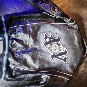May include: A black leather varsity jacket featuring white piping, ribbed cuffs with blue stripes, and large embroidered patches on the back and sleeves. The back displays the letters AV with a circular logo reading Avirex Varsity 1975.