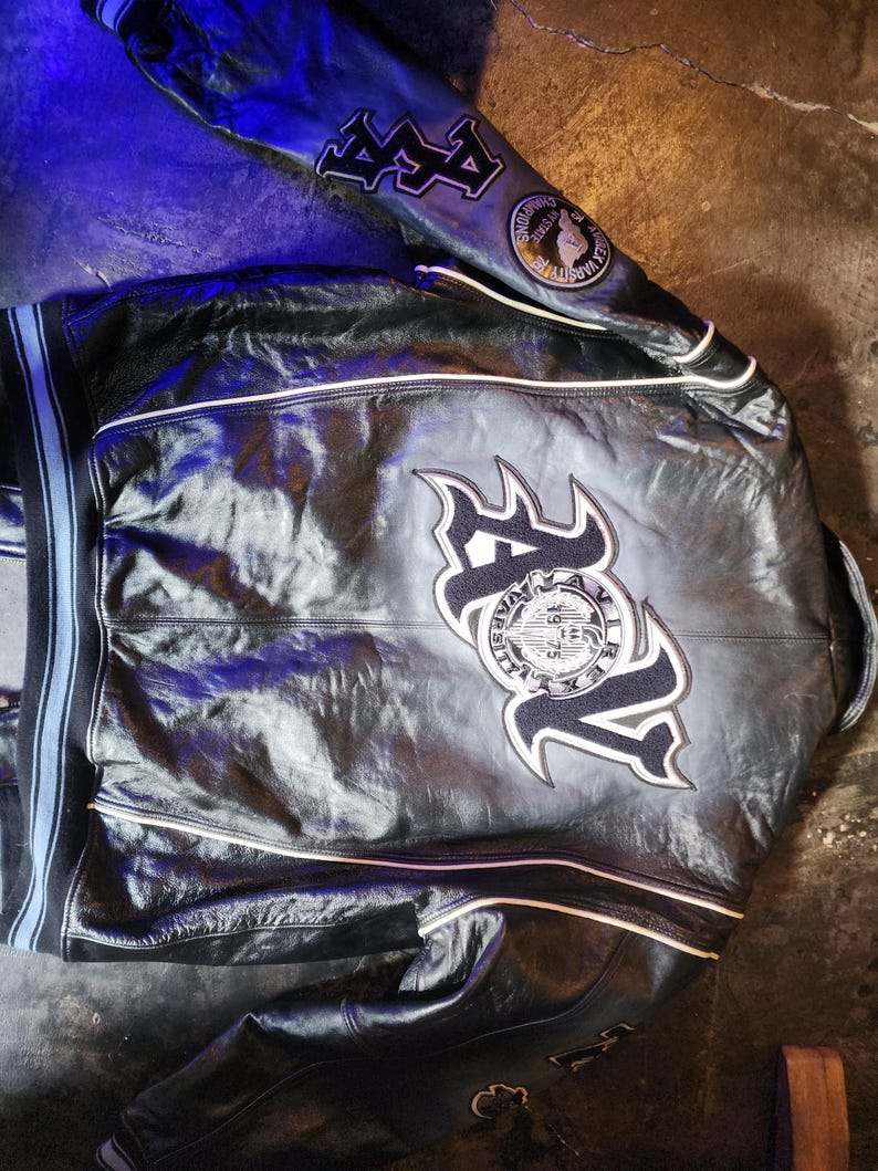May include: A black leather varsity jacket featuring white piping, ribbed cuffs with blue stripes, and large embroidered patches on the back and sleeves. The back displays the letters AV with a circular logo reading Avirex Varsity 1975.