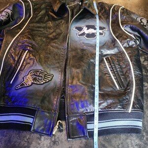 May include: A black leather jacket with white piping, featuring embroidered patches on the chest and sleeves. A measuring tape is placed vertically along the front, indicating a length of approximately 32 inches from the collar to the hem.