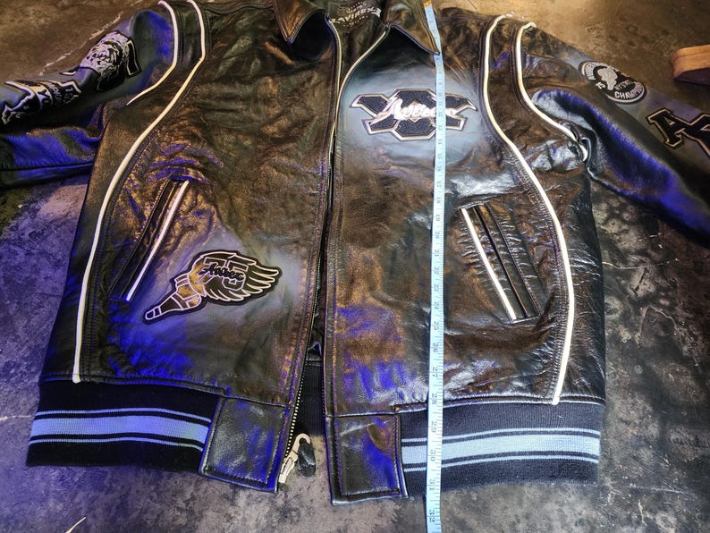 May include: A black leather jacket with white piping, featuring embroidered patches on the chest and sleeves. A measuring tape is placed vertically along the front, indicating a length of approximately 32 inches from the collar to the hem.