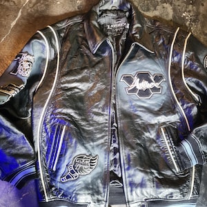 May include: A black leather varsity jacket featuring white piping, a fold-down collar, and a front zipper closure. The jacket displays various embroidered patches, including the text Avirex, a winged 75 logo, and a circular NY State Champions patch.
