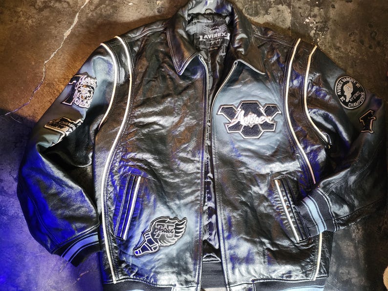 May include: A black leather varsity jacket featuring white piping, a fold-down collar, and a front zipper closure. The jacket displays various embroidered patches, including the text Avirex, a winged 75 logo, and a circular NY State Champions patch.