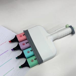 Puede incluir: Una herramienta resaltadora m&uacute;ltiple de pl&aacute;stico blanco con cuatro marcadores Stabilo integrados en tonos pastel rosa, morado, turquesa y verde claro. El dispositivo tiene un marco gris y un mango texturizado, con las puntas alineadas para trazar l&iacute;neas.