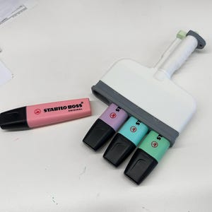 Puede incluir: Un marcador Stabilo Boss Original de color rosa colocado horizontalmente junto a una herramienta de pl&aacute;stico blanca y gris que sostiene tres marcadores Stabilo Boss adicionales en colores pastel morado, azul claro y verde claro.