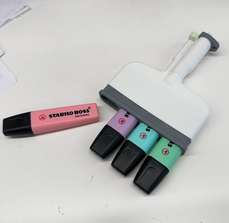 Puede incluir: Un marcador Stabilo Boss Original de color rosa colocado horizontalmente junto a una herramienta de pl&aacute;stico blanca y gris que sostiene tres marcadores Stabilo Boss adicionales en colores pastel morado, azul claro y verde claro.