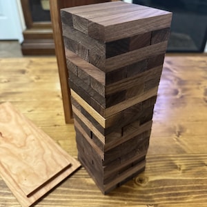 May include: A tall tower of rectangular wooden blocks stacked in alternating horizontal layers. The blocks feature varying shades of brown wood grain. A matching wooden storage box with a separate lid is positioned behind the block tower.