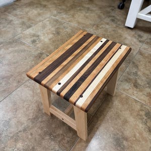 May include: A small wooden stool featuring a rectangular top constructed from alternating strips of dark walnut, medium-toned cherry, and light maple wood. The stool has four sturdy, light-colored wooden legs and visible dark circular joinery.