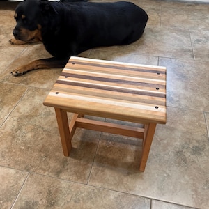 May include: A small wooden stool featuring a rectangular top constructed from alternating strips of light, medium, and dark wood. The top is secured with four visible dark circular fasteners at the corners. The stool has four sturdy legs.