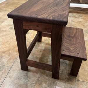 May include: A two-step stool crafted from dark brown wood with a visible grain pattern. The stool features a rectangular top step and a lower step, both supported by four sturdy legs with visible joinery details on the sides and top surfaces.