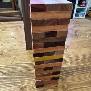 May include: A vertical tower constructed from rectangular wooden blocks of varying natural wood tones, including light, medium, and dark brown shades, with one distinct yellow block integrated into the stack, arranged in a classic block-stacking game formation.