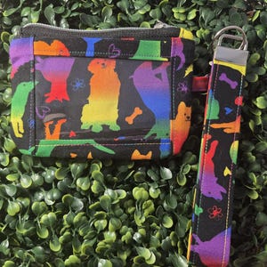 May include: A black zippered pouch and a matching wristlet strap featuring a vibrant rainbow-colored pattern of dog silhouettes, bones, and small hearts. The fabric has a repeating print of various canine shapes in gradient colors against a black background.