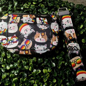 May include: A rectangular black fabric pouch featuring a repeating pattern of cartoon cat faces wearing rainbow-colored glasses and accessories, accompanied by a matching fabric wrist strap attached with a silver-toned metal clasp and hardware.