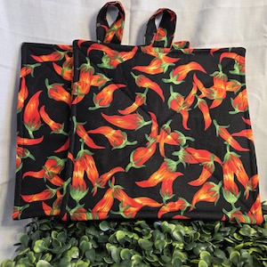 Puede incluir: Dos agarraderas negras acolchadas con un vibrante estampado de chiles rojos y naranjas y hojas verdes. Cada agarradera tiene un lazo de tela a juego para colgar. El fondo es de color claro y neutro.