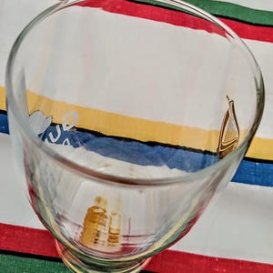 May include: A clear glass goblet with a wide bowl and a short stem. The glass has a gold harp design and other gold graphics. The goblet is sitting on a colorful striped surface with red, green, blue, and yellow stripes.