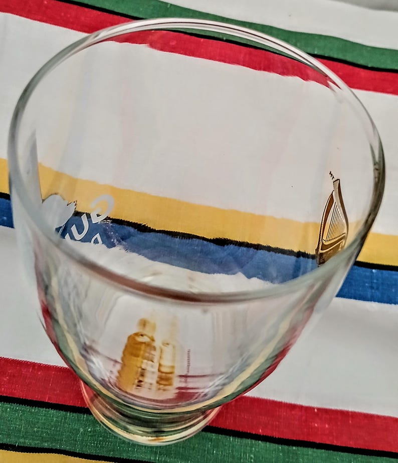 May include: A clear glass goblet with a wide bowl and a short stem. The glass has a gold harp design and other gold graphics. The goblet is sitting on a colorful striped surface with red, green, blue, and yellow stripes.