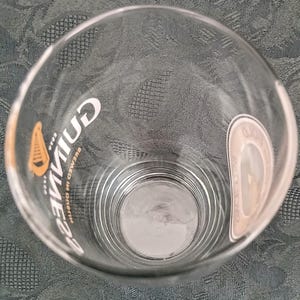 Może przedstawiać: Przezroczysty szklany kufel z logo Guinness w kolorze białym i złotym. Szklanka ma zakrzywiony kształt i grubą podstawę. Logo Guinness jest wydrukowane pionowo z boku. Szklanka jest pusta.
