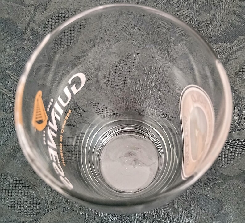 Może przedstawiać: Przezroczysty szklany kufel z logo Guinness w kolorze białym i złotym. Szklanka ma zakrzywiony kształt i grubą podstawę. Logo Guinness jest wydrukowane pionowo z boku. Szklanka jest pusta.