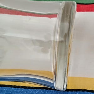 May include: A clear, rectangular glass dish with a curved side. The dish reflects the colors of the striped fabric beneath it, including red, white, yellow, and blue. The glass has a smooth, polished appearance.
