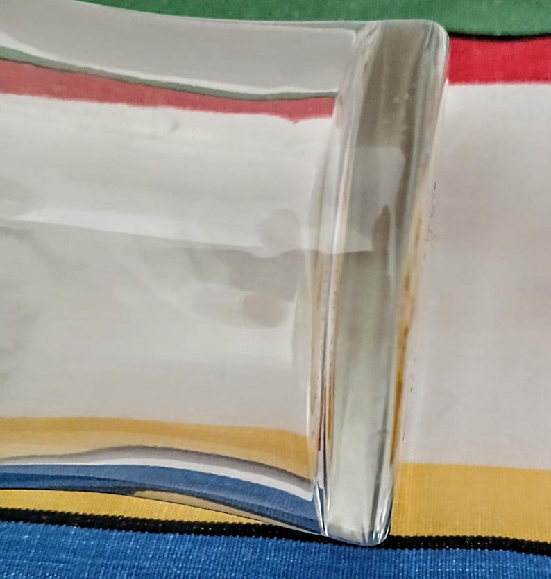 May include: A clear, rectangular glass dish with a curved side. The dish reflects the colors of the striped fabric beneath it, including red, white, yellow, and blue. The glass has a smooth, polished appearance.