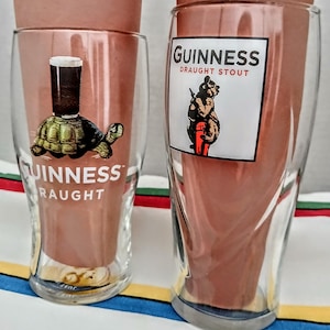 May include: Two clear Guinness pint glasses. One glass features a turtle with a pint of stout on its back, and the word "Guinness" and "Draught" printed below. The other glass has a bear holding a pint of stout, with the words "Guinness Draught Stout".