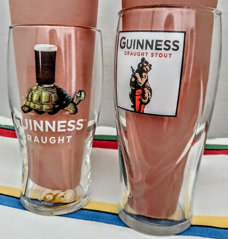 May include: Two clear Guinness pint glasses. One glass features a turtle with a pint of stout on its back, and the word "Guinness" and "Draught" printed below. The other glass has a bear holding a pint of stout, with the words "Guinness Draught Stout".